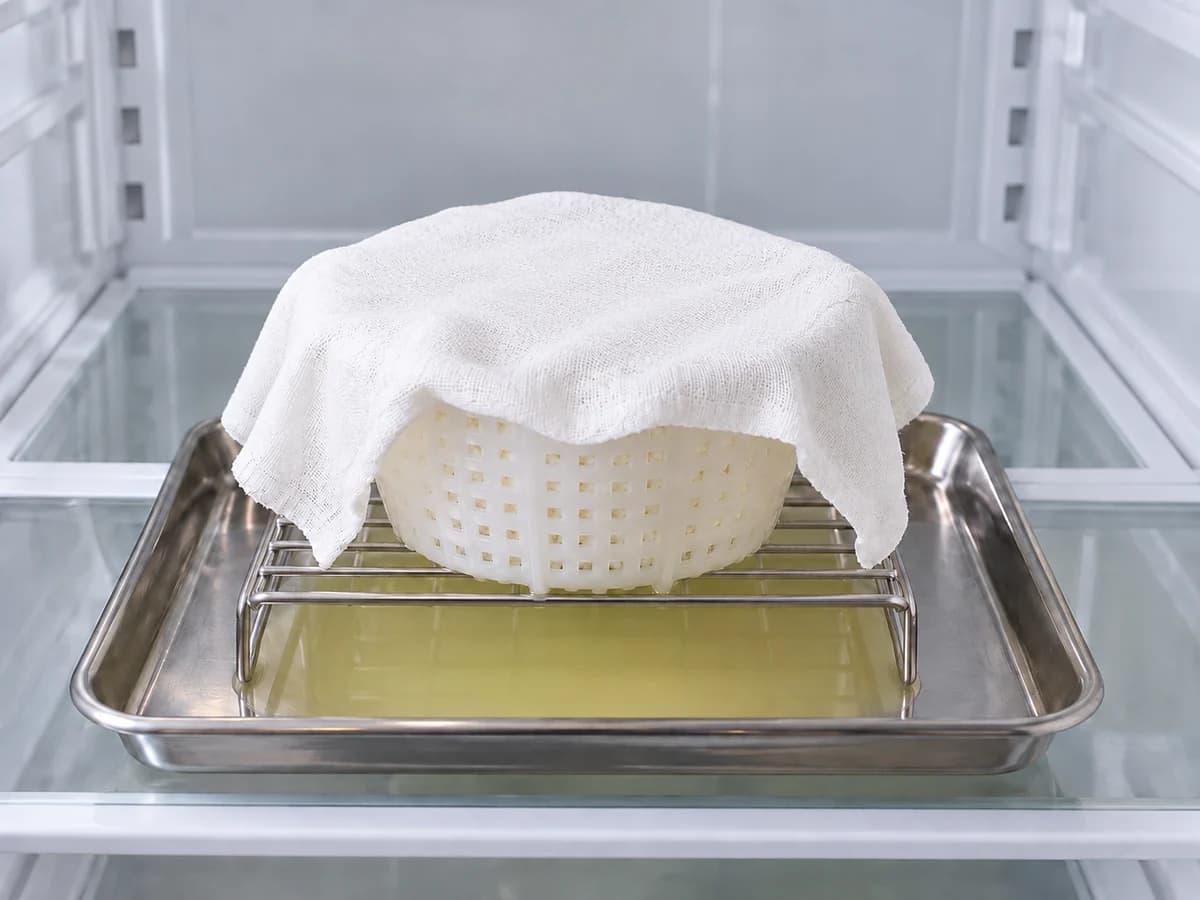 Covered cheese mold raised above collected whey in a clean refrigerator
